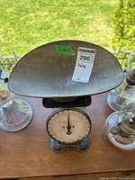 Front top-down view showing metal curved weighing pan, dial with measurement pointer and surrounding glass oil lamps on a wooden table with a green yard background.