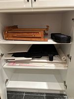 Two shelves inside white cabinet with foldable wooden easel folded and stacked art paper, and black art carry bag