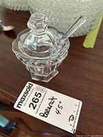 Clear Baccarat crystal mustard pot with lid and matching spoon displayed on a wooden surface next to a stack of clear glass plates and other kitchen items.