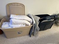 Two plastic storage bins side by side, the beige bin open with white mattress covers inside, and the dark gray bin next to it with some clear bags leaning on it.