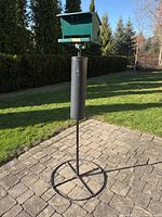 Full view of the bird feeder on metal pole with circular base on paved outdoor surface, greenery in background