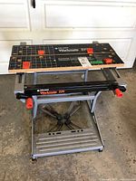 Front view of the Black and Decker Workmate 225 folding work bench showing the work surface with measurement guides, the metal frame base, and orange clamps and handles.