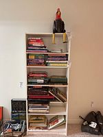 A white shelving unit with six shelves filled with books and a sitting rooster figurine on top.