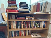 Wide shot of wooden shelf with books in various sizes, some stacked on top and some arranged side by side. Includes fiction, non-fiction, and some reference books like histories and dictionaries.