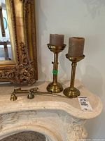 Photo showing two brass candleholders with brown pillar candles seated on a light-colored surface near an ornate gold frame mirror. Also includes two brass candle snuffers laid out in front of the candleholders.