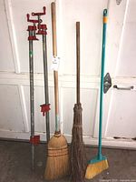 Two red metal pipe clamps and three brooms including a corn broom, reed broom, and plastic broom standing against the garage door.