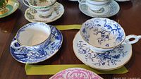 Two blue and white Coalport teacups and matching saucers displayed on a wooden table with other china items in background, showing intricate scenic patterns with gold trim.