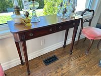 Wooden console table positioned in front of window, showing front and right side including drawer details and metal ring pulls. Surface has minor wear.