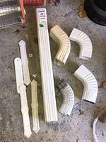 White eavestrough parts including elbows and straight downspout sections arranged on floor, showing size and condition.