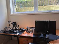 Image of all electronics on an office desk by the window, including desk phone, desk lamps, cordless phones, Acer monitor, and cables.