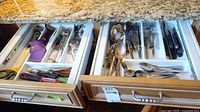 Two wooden drawers open showing organized cutlery and utensils in white plastic trays on granite countertop. Items include forks, knives, spoons, scissors, and other kitchen utensils.