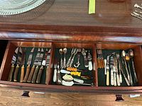 Drawer with organized compartments containing knives with wooden handles, silver plate butter knives, spoons, and other small utensils.