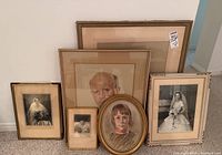 Six framed portraits arranged on carpeted floor against wall, various sizes and frame styles