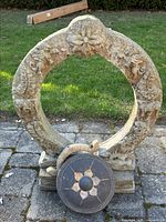 Front view of large circular concrete garden ornament with floral and patterned carvings and a separate round decorative concrete plate with central knob and flower design set in front.