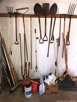 Overview of long-handled tools hung on wall rack and items on floor including rakes, shovels, spades, spray containers, fuel cans and broom