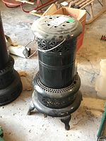 Front angled view of Valor metal kerosene heater showing detailed metal ventilation design, handle and base with 3 decorative legs on concrete garage floor with other tools and boxes in background.