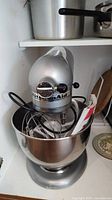 Front view of silver KitchenAid Custom stand mixer fitted with stainless steel bowl and wire whisk on lower cabinet shelf