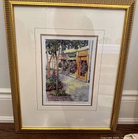 Framed limited edition print showing a colorful street scene with storefronts, trees, and flowers. The print is matted and housed in a decorative gold frame.