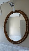 Full view of oval gold framed mirror hanging on wall under light fixtures, showing overall size and shape.