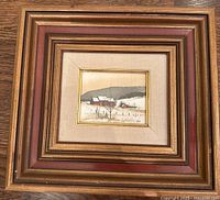 Front view of the small original painting showing the red barn and snow scene, within ornate gold and reddish wooden frame and beige mat.