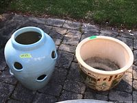 Two ceramic planters shown outdoors on stone paving, blue strawberry planter with circular holes on the left and beige decorative planter on the right.