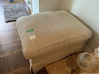 Front angled view of beige upholstered ottoman showing cushion top and skirt base with floor vent visible nearby