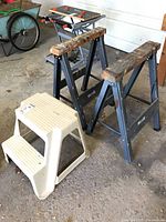 Lot containing two metal saw horses with wooden tops and a plastic two-step stool, placed in garage floor area showing concrete and surrounding items.