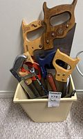 Overview of assorted saws placed in a plastic container showing various types of saws with wooden, metal, and plastic handles including crosscut saws and hacksaws.