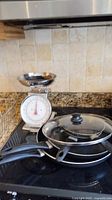 Full view of all items on stove including kitchen scale and three frying pans with glass lid on top