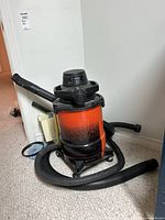 Full view of the orange and black 5 gallon Shop Vac with attached hose and nozzle attachments coiled around it on the floor.
