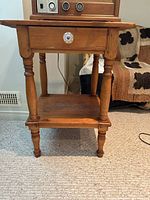 Front view of wooden side table with one drawer and lower shelf, showing turned legs and white knob on drawer.