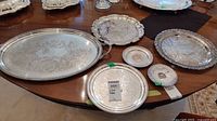 Photo of multiple silver-plated trays and platters arranged on table showing detailed engraving and overall condition with wear and tarnish.