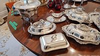 Photo showing the assortment of silver plated serving pieces arranged on table, including lidded chafing dish, gravy boat, two covered rectangular serving dishes, and tray.