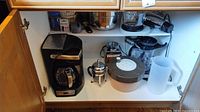 Photo of cabinet shelf with Delonghi coffee maker, French press, blender base, and other kitchen items inside cabinet.