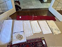 Top-down view of multiple folded white and cream table linens including lace, embroidery, and scalloped edges laid out near a red food warmer mat with attached electrical cord.