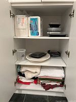 White cabinet with three shelves containing kitchen towels, ceramic platter, serviettes, napkins, and plastic containers.