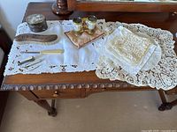 Photo shows all items arranged on wooden table covered with embroidered white linen; includes opera glasses, brushes, metal box, and linens