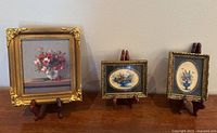 Three framed floral art pieces displayed with wooden stands on a surface against a wall.