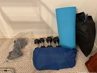 Four pairs of hex rubber dumbbells with labeled weights, yoga mat rolled up and standing vertically, dark blue sleeping bag in storage bag, and black packed air mattress.