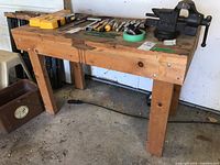 Wood workbench with attached metal vise shown from left side angle. The bench shows signs of wear and has various tools placed on top which are not included.