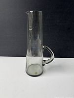 Clear glass decanter with tall cylindrical shape, angular handle on one side, and narrow spout for pouring. Displayed against black background.
