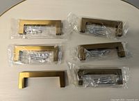 Top view of six brass drawer handles individually packaged with screws, arranged in two rows of three on a light surface.