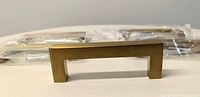 Single brass drawer handle with rectangular shape and flat grip, shown unpackaged with packaged handles in background.
