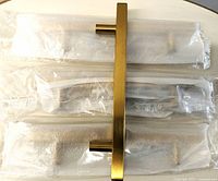 Set of 5 brass drawer pulls shown individually wrapped in plastic packaging with one pull unwrapped for display showing a polished brass finish and modern bar handle design.