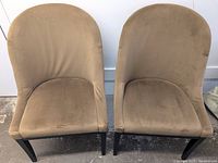 Front view of two light brown curved back upholstered chairs, showing soft fabric upholstery and nailhead trim. Some marks visible on one seat.