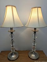 Two silver metal lamps with three clear acrylic spherical accents each and cream bell-shaped shades on a wooden surface, lit to show light diffusion.