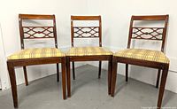 Three vintage wooden dining chairs are shown side by side in a plain setting, highlighting their decorative backrest design and plaid upholstered seat.