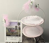Photo of white metal two-tiered dessert stand holding two scalloped paper plates, decorated with pink and white tulle and artificial white roses, next to a white metal picture frame with sample image.