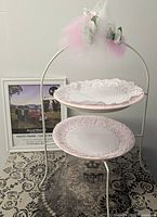 Photo of two tiered white metal dessert stand with two decorative paper plates and white metal picture frame in background