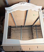 Full view of the white wooden framed mirror resting horizontally, showing the entire rectangular mirror with the carved applique decorations at the top center and corners.